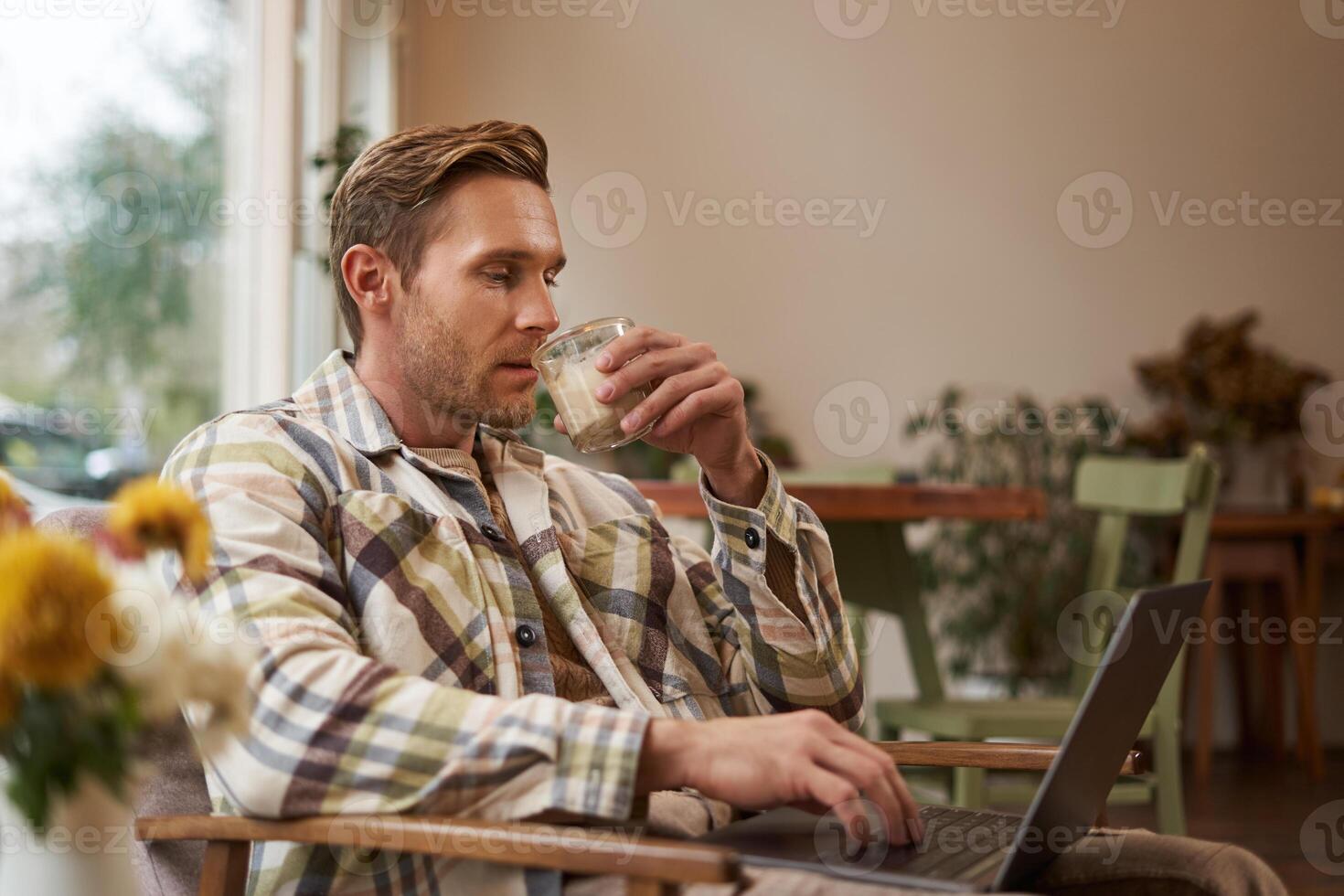 Portrait of handsome young man, working in cafe, sitting with laptop and coffee in hand, has remote job and using local co-working space instead of going to the office photo