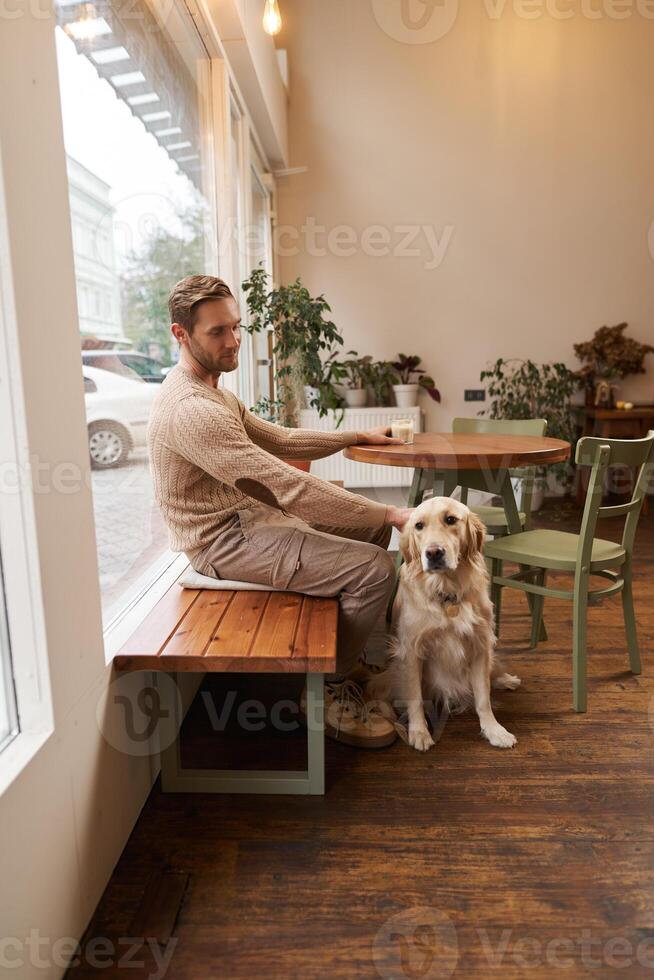 Vertical shot of young man, owner of golden retriever, sitting in cafe with and stroking his dog under the table, drinking coffee in a coffeeshop photo