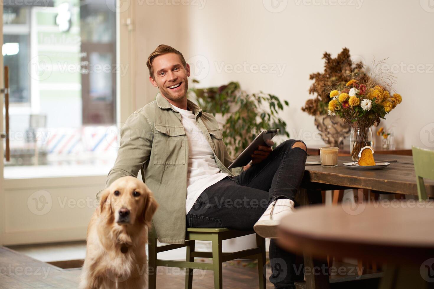 Portrait of handsome young man, cafe visitor, sitting with his dog, petting golden retriever and reading news on digital tablet, concept of lifestyle and pet-friendly public places photo