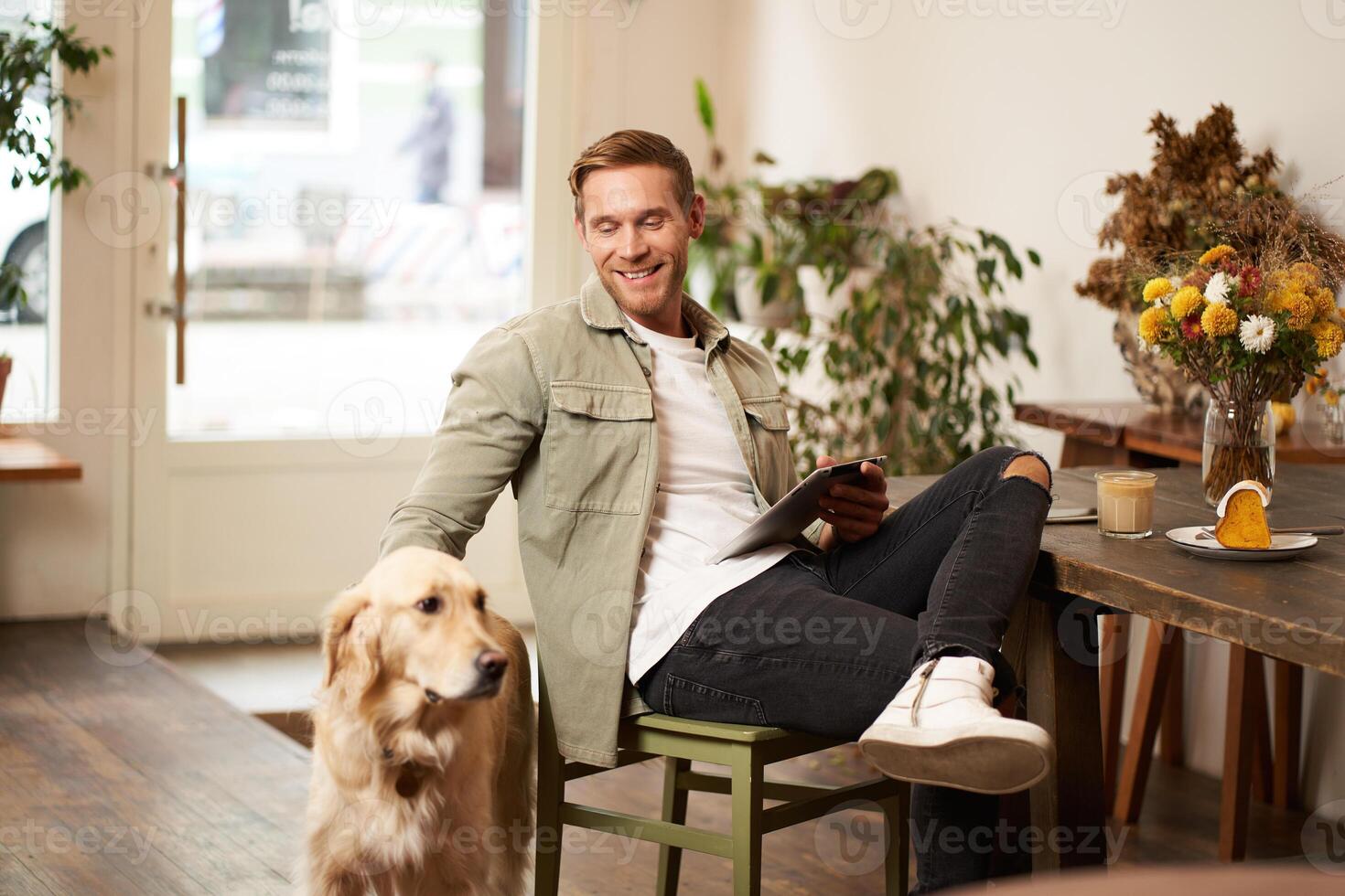 Portrait of happy smiling young man, cafe visitor, sitting in coffee shop with his dog, petting golden retriever, holding digital tablet, reading news or browsing on internet photo