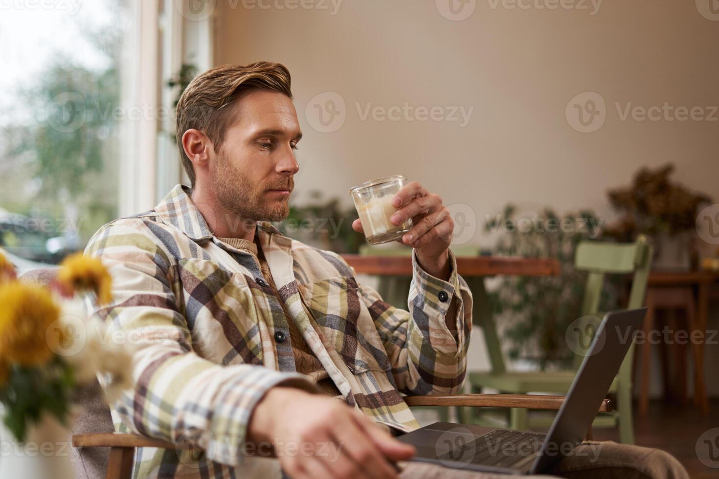 Portrait of young freelancer, a man in casual clothes, sitting in cafe and drinking coffee, working remotely on laptop, digital nomad doing his project outdoors in co-working space photo