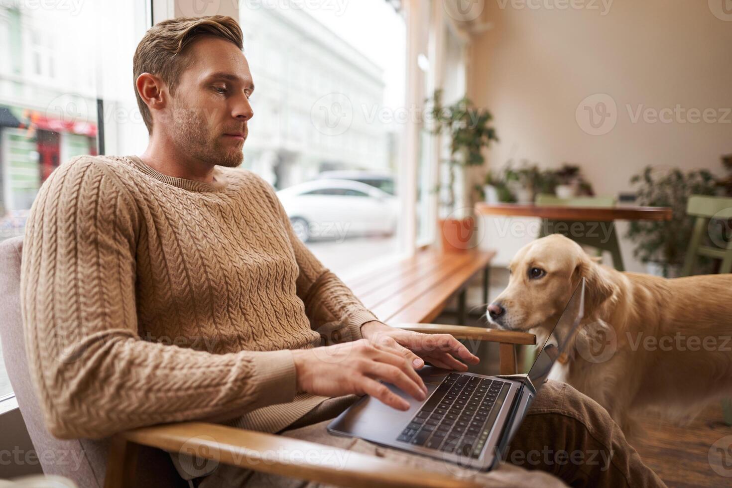 Portrait of young working man sitting in cafe, typing on keyboard while a dog looking at him. Concentrated coffee shop visitor doing his job online, sitting near window in co-working space photo