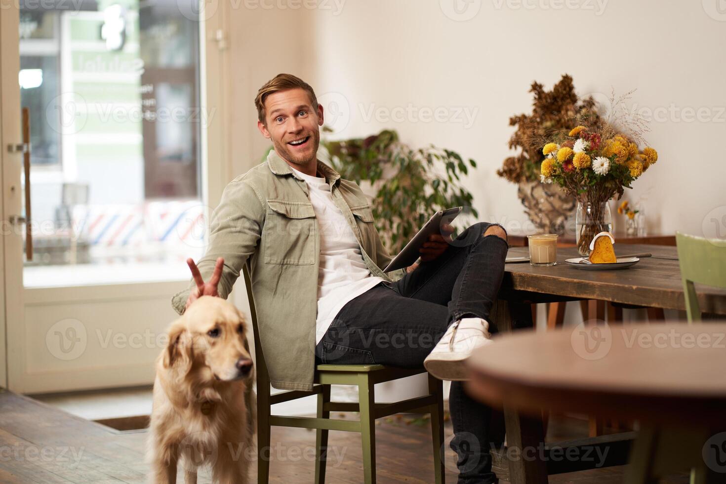 Portrait of happy smiling young man, cafe visitor, sitting in coffee shop with his dog, petting golden retriever, holding digital tablet, reading news or browsing on internet photo