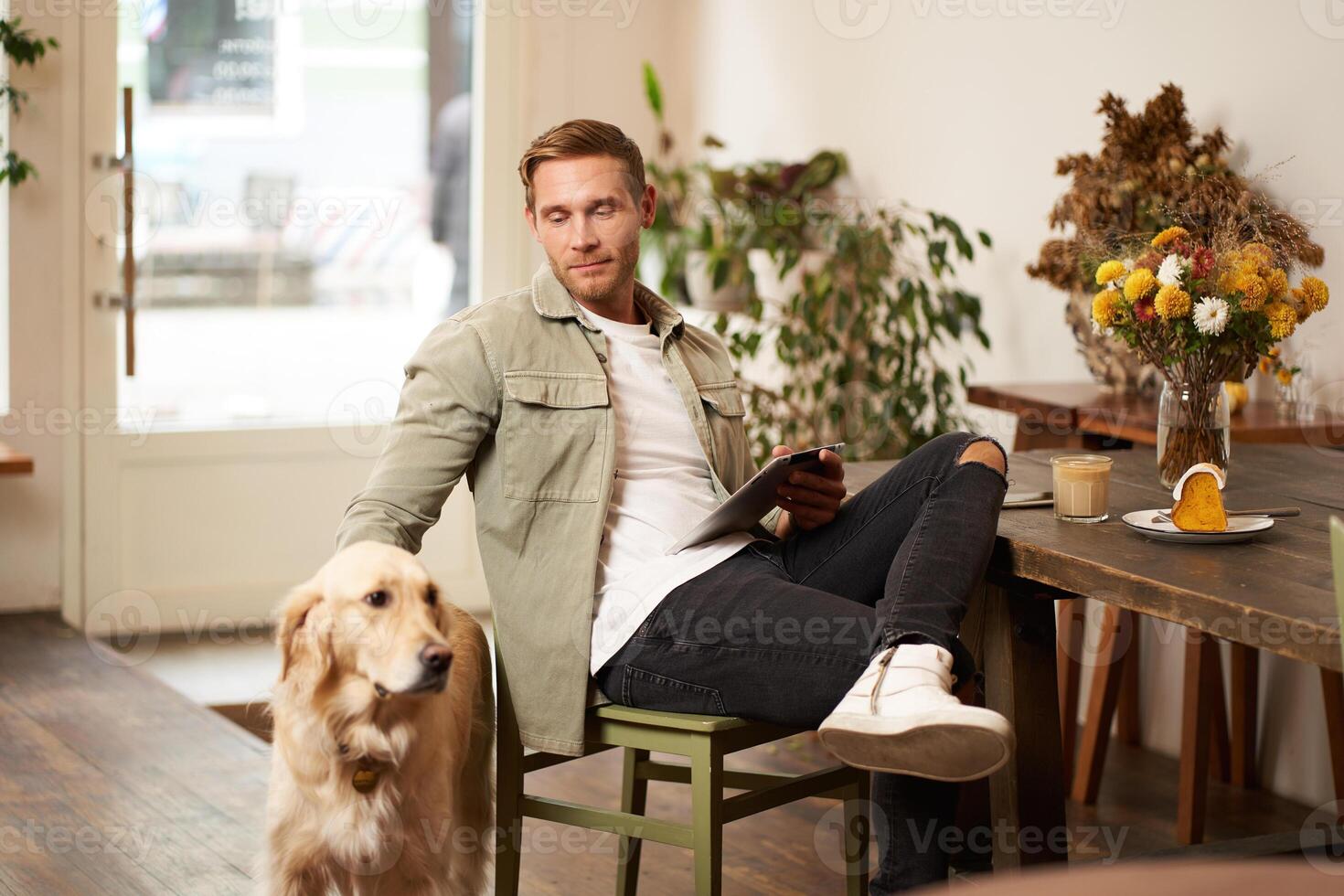 Portrait of handsome young man sits in cafe with his dog, petting golden retriever, reading news on tablet, relaxing with cup of coffee photo