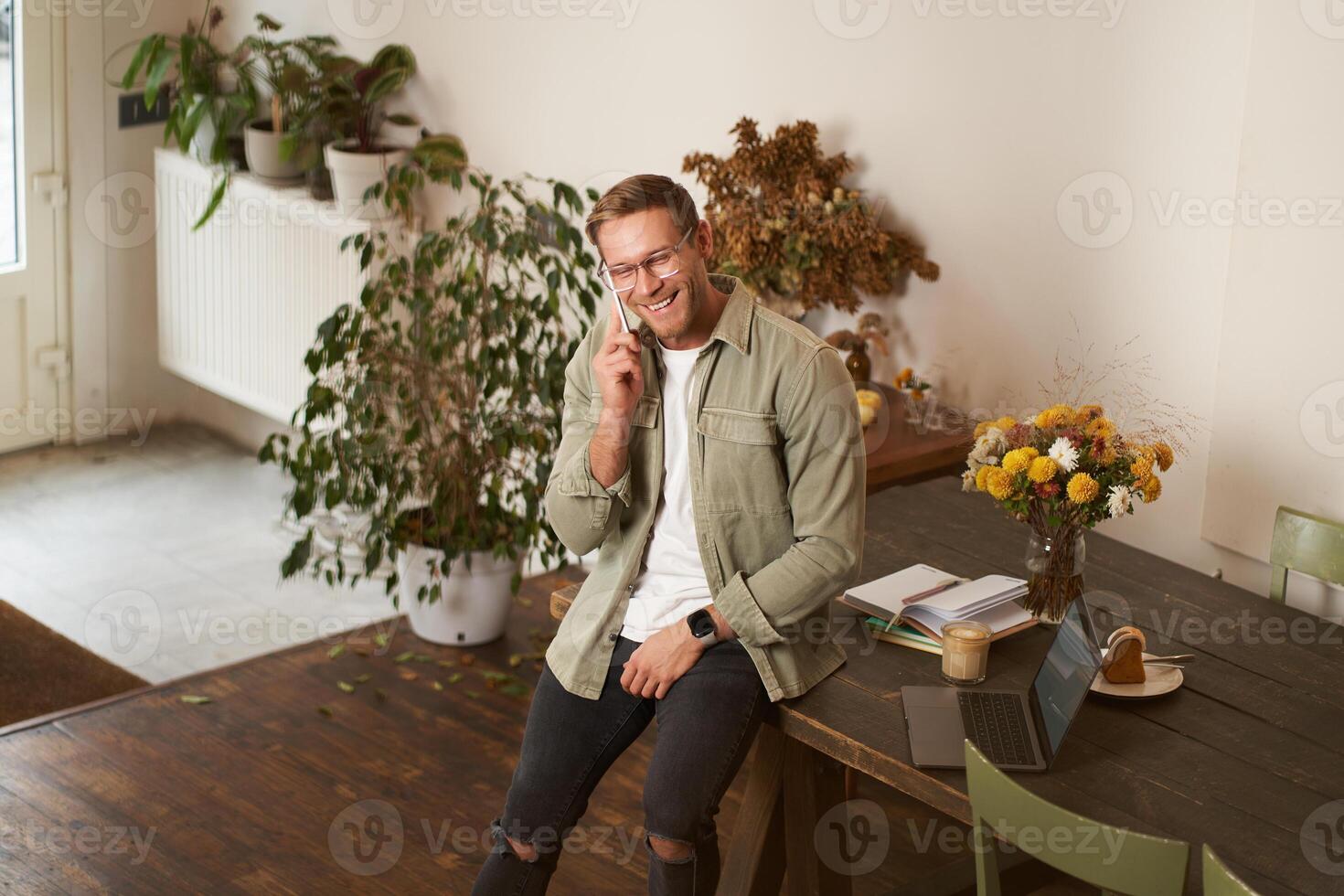 Portrait of handsome office manager, guy in co-working space, talking on mobile phone, calling someone, laughing and smiling, having pleasant conversation, sitting in office on table photo