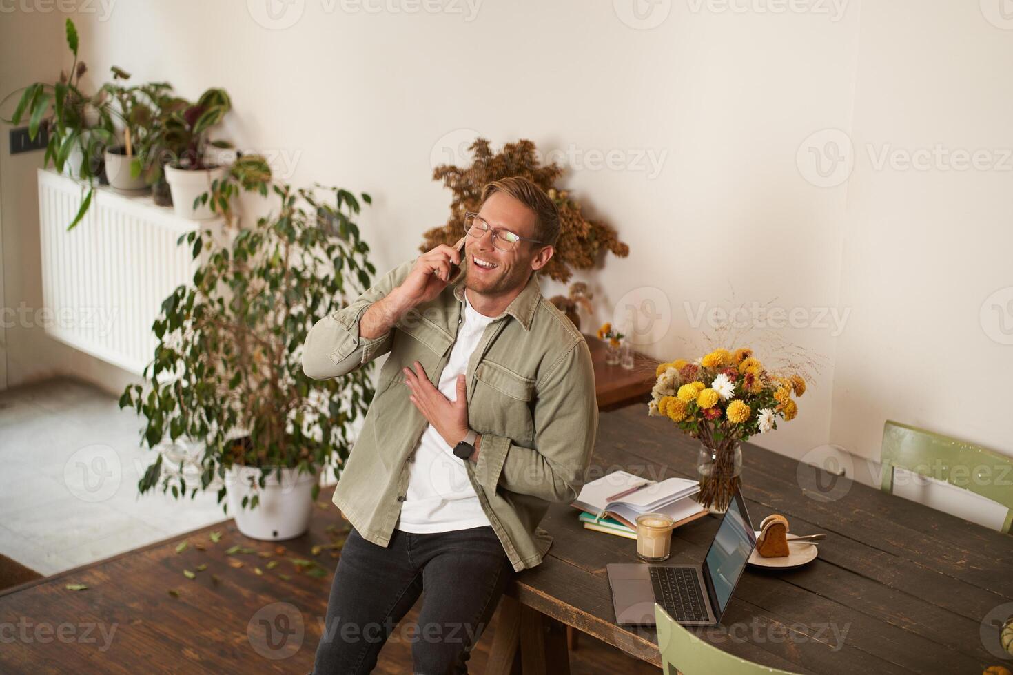 Portrait of handsome office manager, guy in co-working space, talking on mobile phone, calling someone, laughing and smiling, having pleasant conversation, sitting in office on table photo