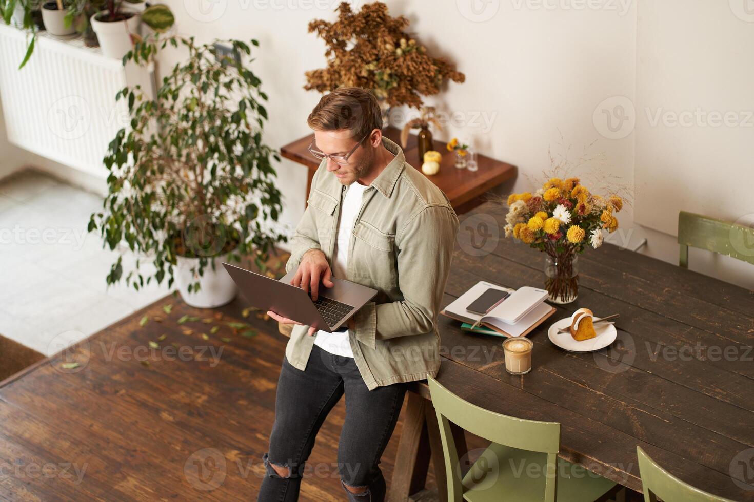 Handsome manager, man in glasses sits on table, holds laptop and types, sends message, works on project, drinks coffee, spends time at co-working space or an office photo
