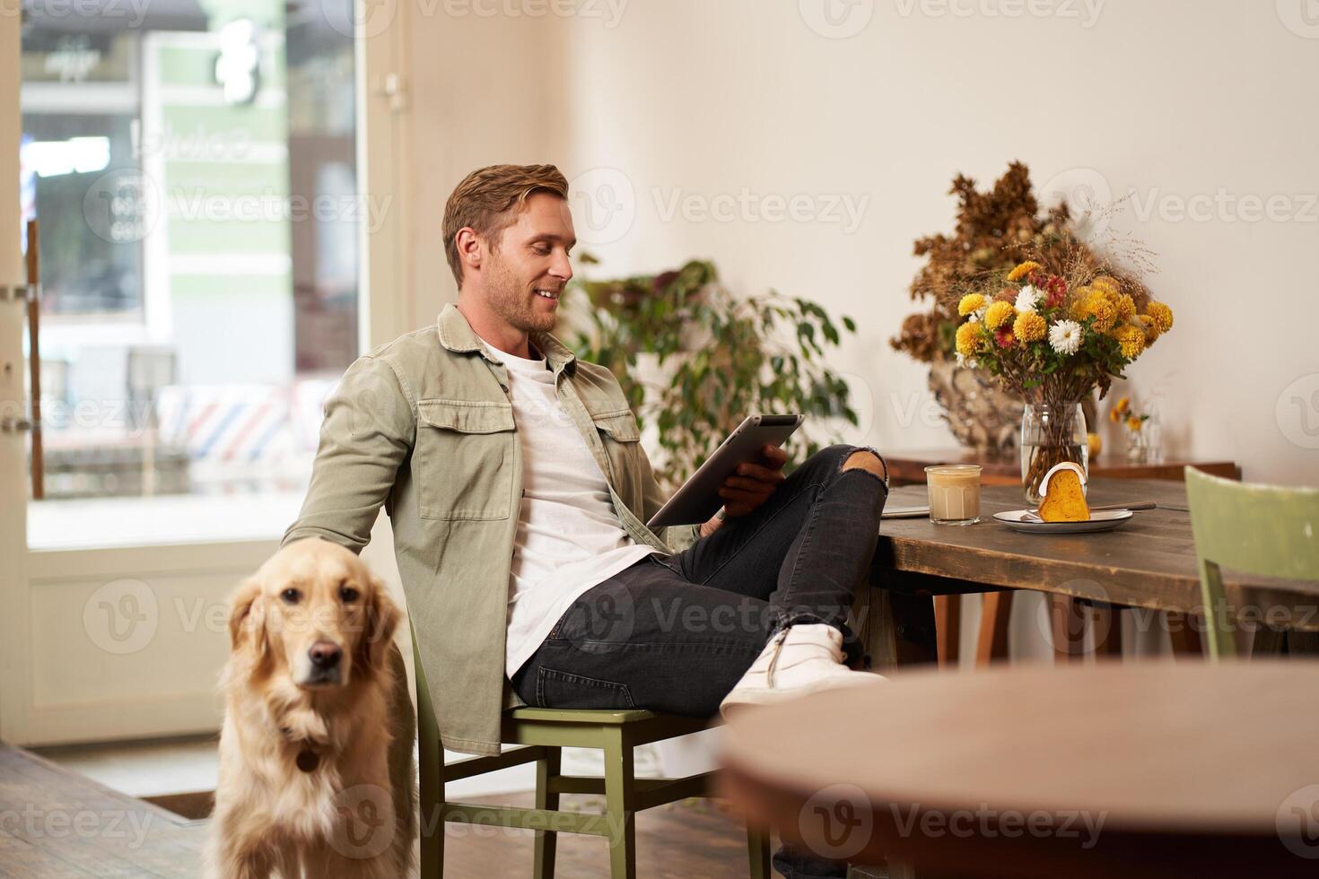 Portrait of handsome young man sits in cafe with his dog, petting golden retriever, reading news on tablet, relaxing with cup of coffee photo