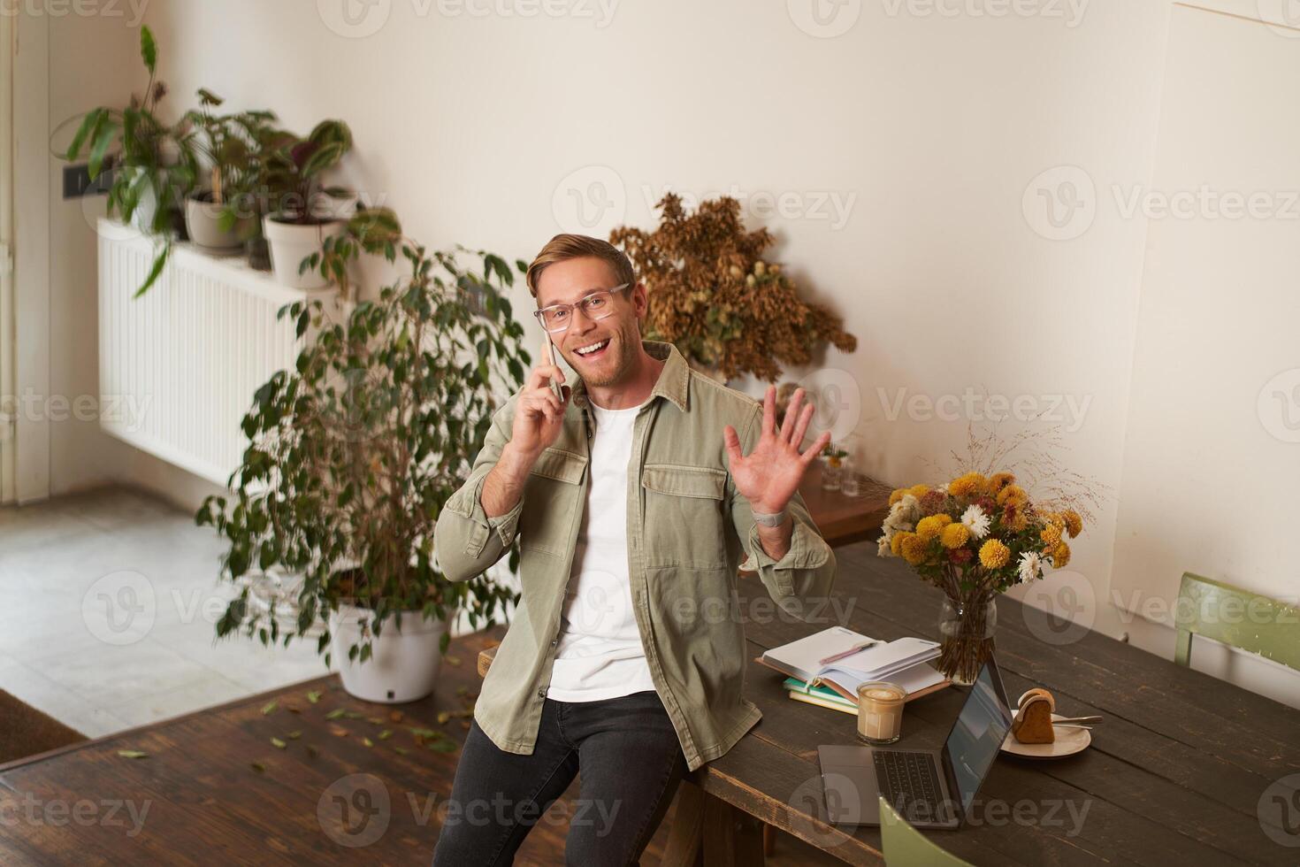 Handsome smiling young man in glasses, sits on office table, waves hand to say hello to you, looking at camera, talks on mobile phone, makes a telephone call photo