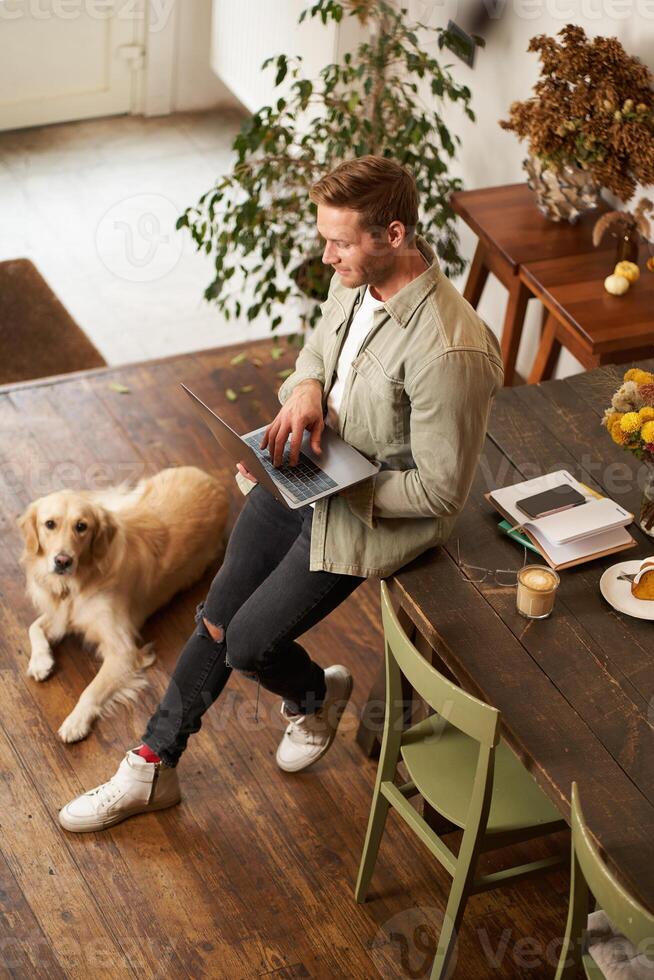 Vertical shot of handsome young man with a dog, leaning on table, holding laptop in hands, looking at screen, checking emails at work. Businessman at co-working space with his golden retriever photo
