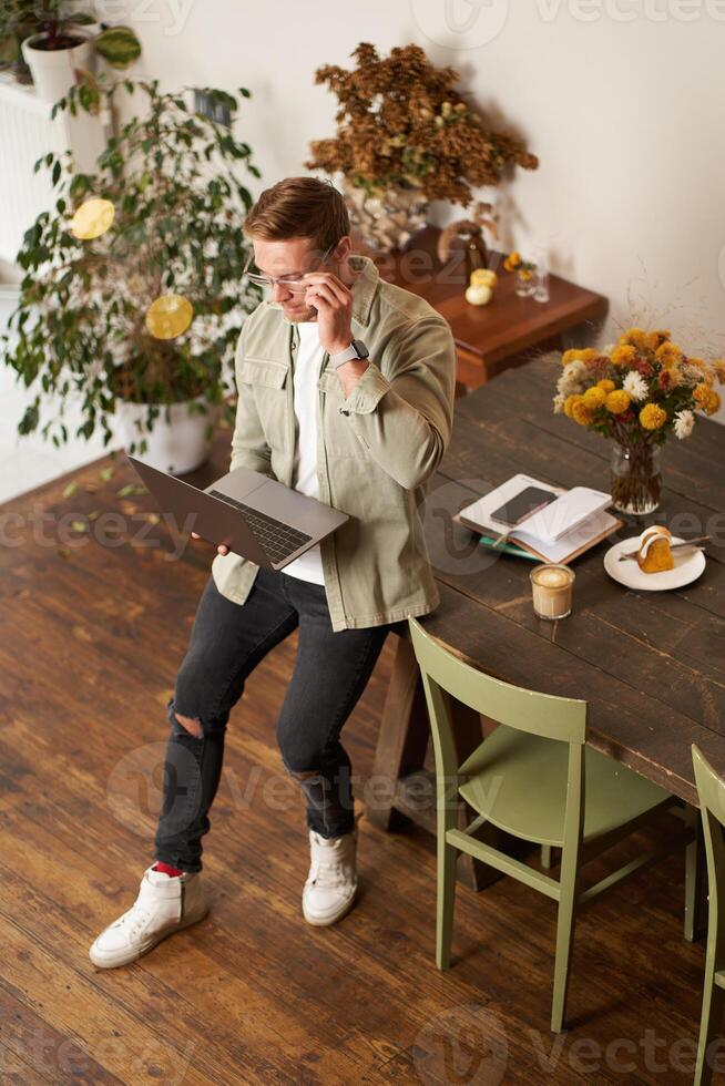 Portrait of handsome young man sits with laptop, looks at screen with concentrated, thoughtful face, working on a project, e-learning at co-working space, drinking coffee photo