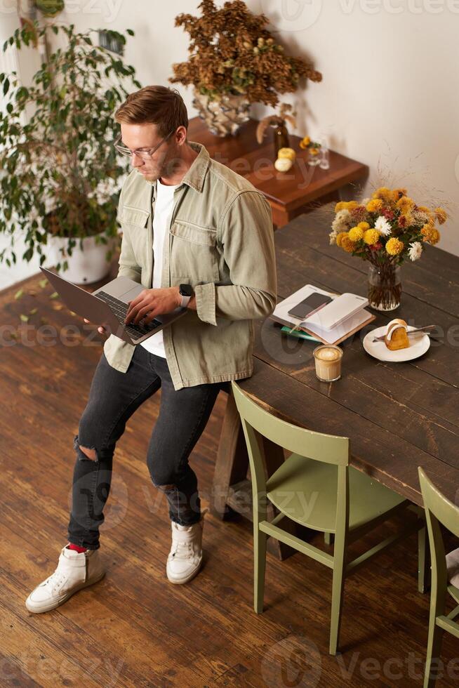Portrait of handsome young man sits with laptop, looks at screen with concentrated, thoughtful face, working on a project, e-learning at co-working space, drinking coffee photo