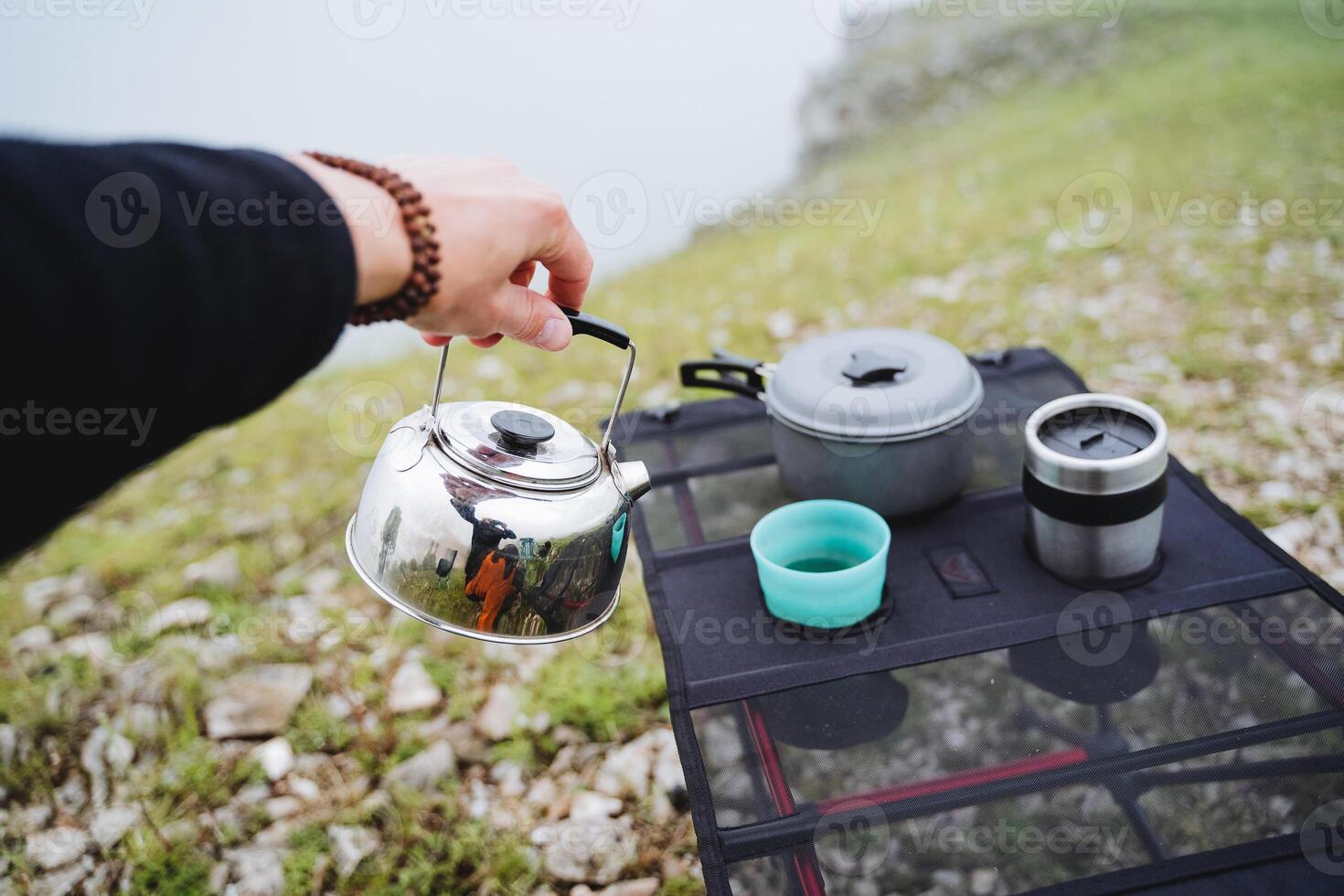 desayuno de un turista en un caminata sentado en el montaña a el mesa, mano brebajes té hirviendo agua en un taza, un metal pava, tomar un mano por el manejar, dos anteojos, un pan para comiendo foto