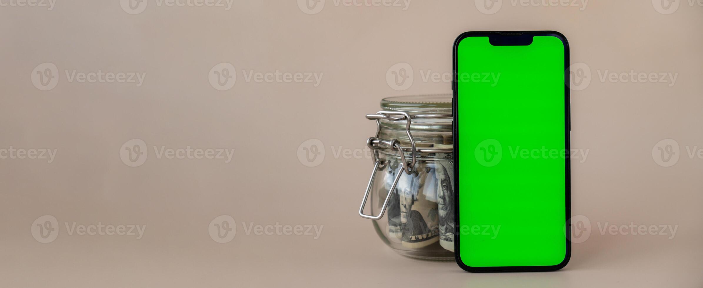 Banner Vertical Green screen on modern mobile phone in background of glass jar full of American currency dollar banknotes on beige background. Cope space for text. Advertisement for application website. Concept of money economy banks and finances photo