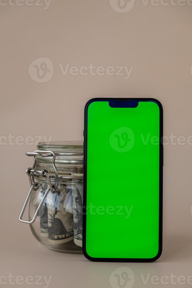 Vertical Green screen on modern mobile phone in background of glass jar full of American currency dollar banknotes on beige background. Cope space for text. Advertisement for application website. Concept of money economy banks and finances photo