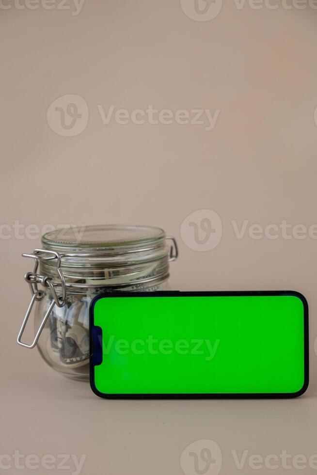 Chrome key smartphone screen mock up template in horizontal position on beige background. Copy space App website advertising. Jar filled with dollars cash. Concept of Mobile application and technology business savings. photo