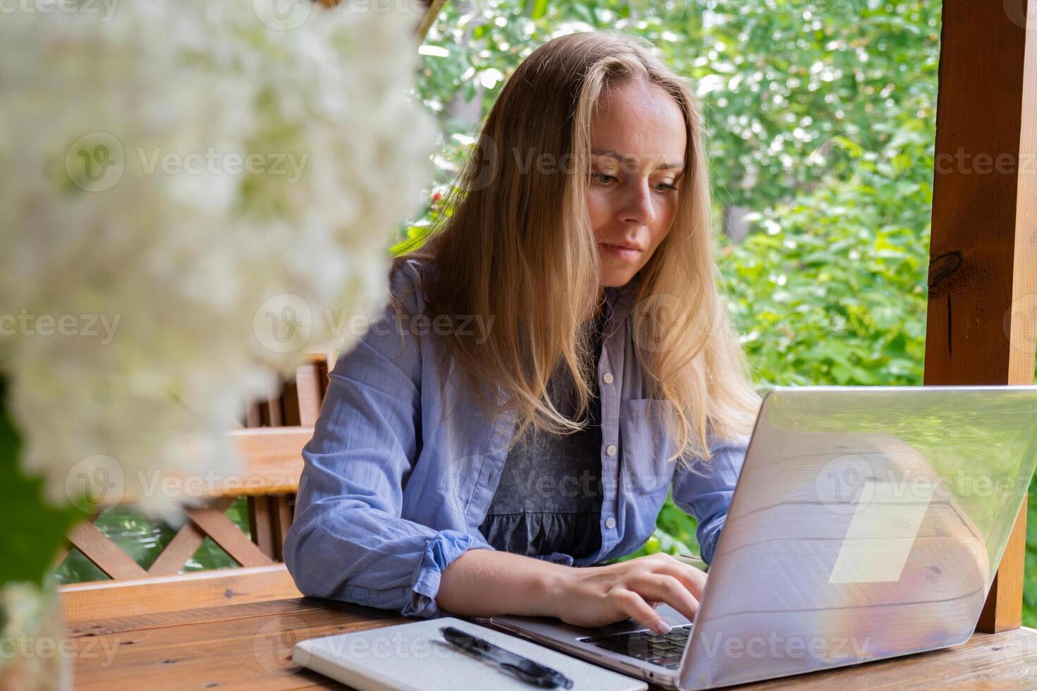 hembra estudiante tiene en línea lección educación al aire libre en jardín de madera hueco. rubia mujer sentado fuera de trabajo en ordenador portátil teniendo llamar. unidad con naturaleza foto