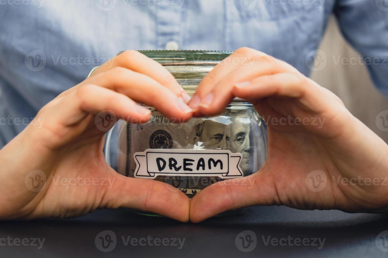 Unrecognizable woman showing heart sign Saving Money In Glass Jar filled with Dollars banknotes. DREAM transcription in front of jar. Managing personal finances extra income for future insecurity background photo