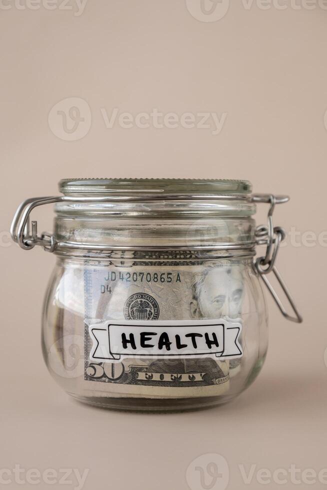 Saving Money In Glass Jar filled with Dollars banknotes. HEALTH transcription in front of jar. Managing personal finances extra income for future insecurity. Beige background photo