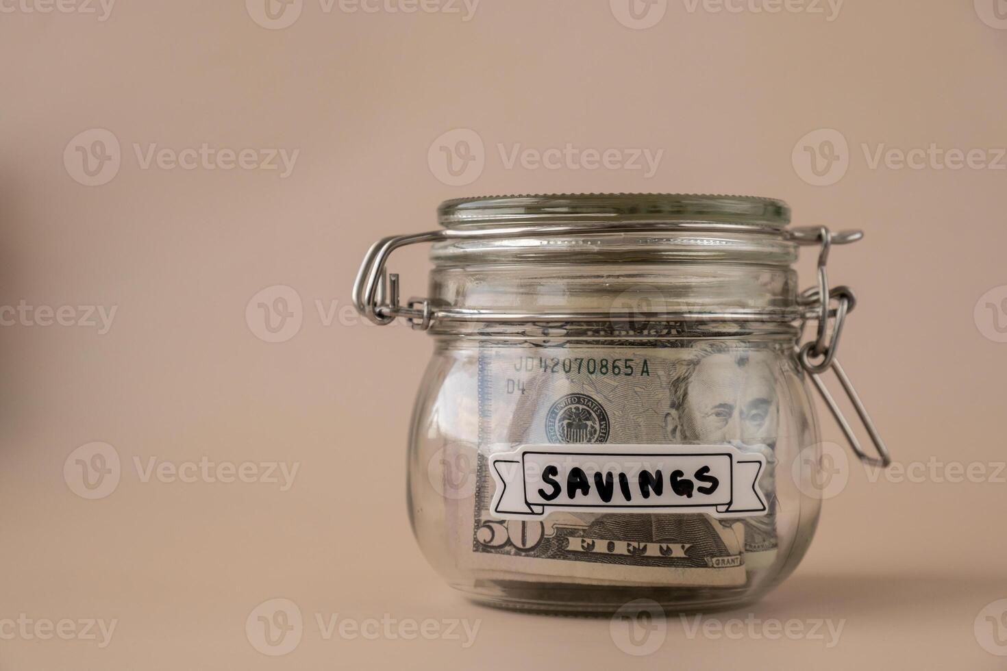 Saving Money In Glass Jar filled with Dollars banknotes. SAVINGS transcription in front of jar. Managing personal finances extra income for future insecurity. Beige background photo