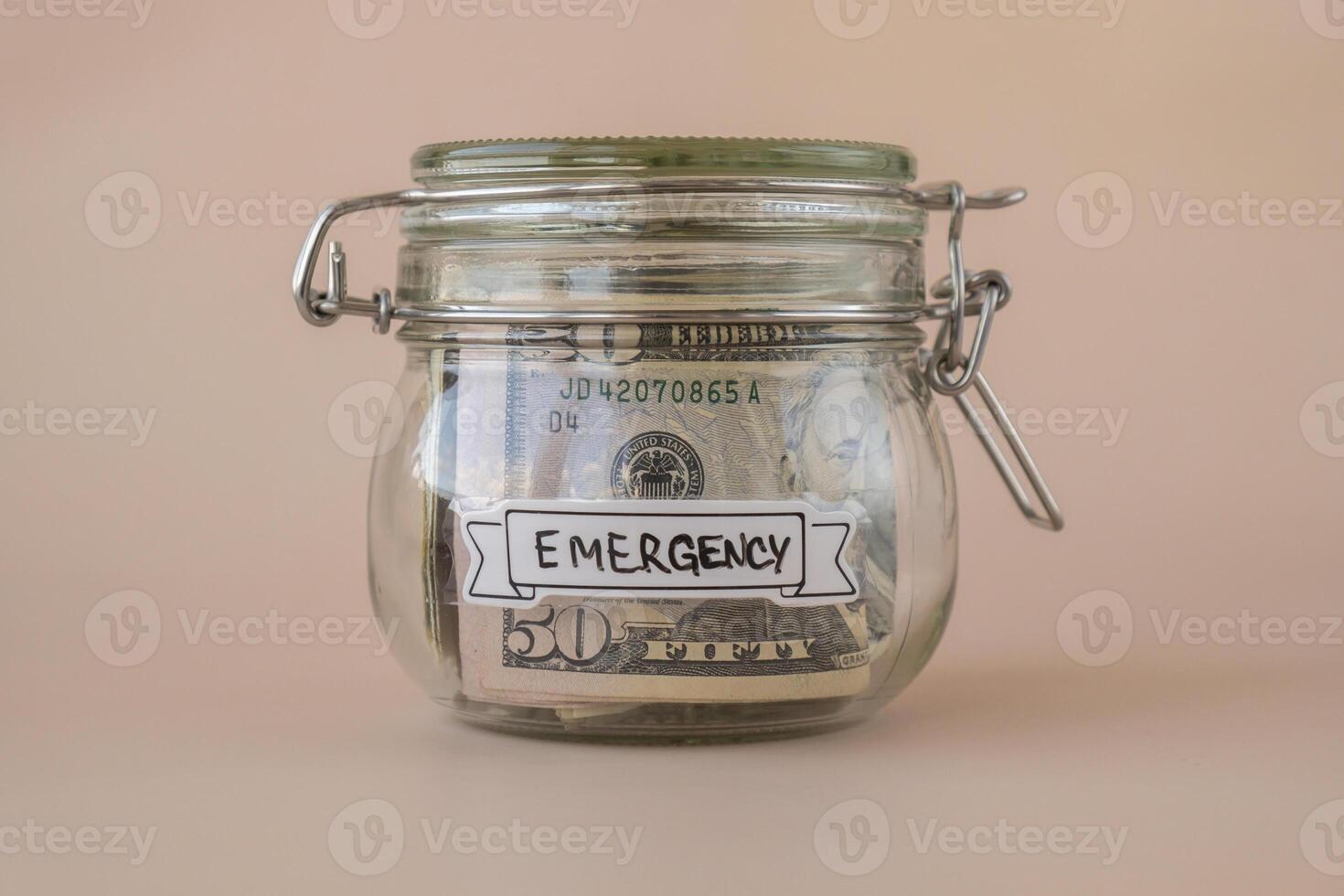 Saving Money In Glass Jar filled with Dollars banknotes. EMERGENCY transcription in front of jar. Managing personal finances extra income for future insecurity. Beige background photo