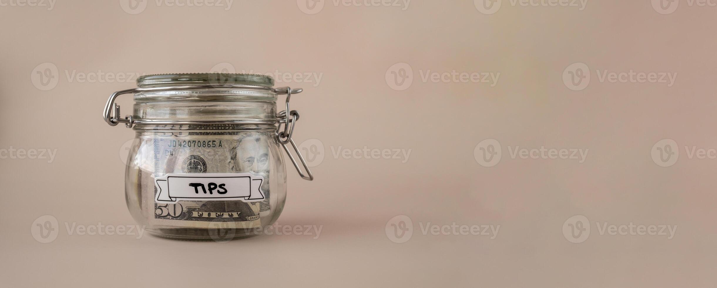 Saving Money In Glass Jar filled with Dollars banknotes. TIPS transcription in front of jar. Managing personal finances extra income for future insecurity. Beige background photo