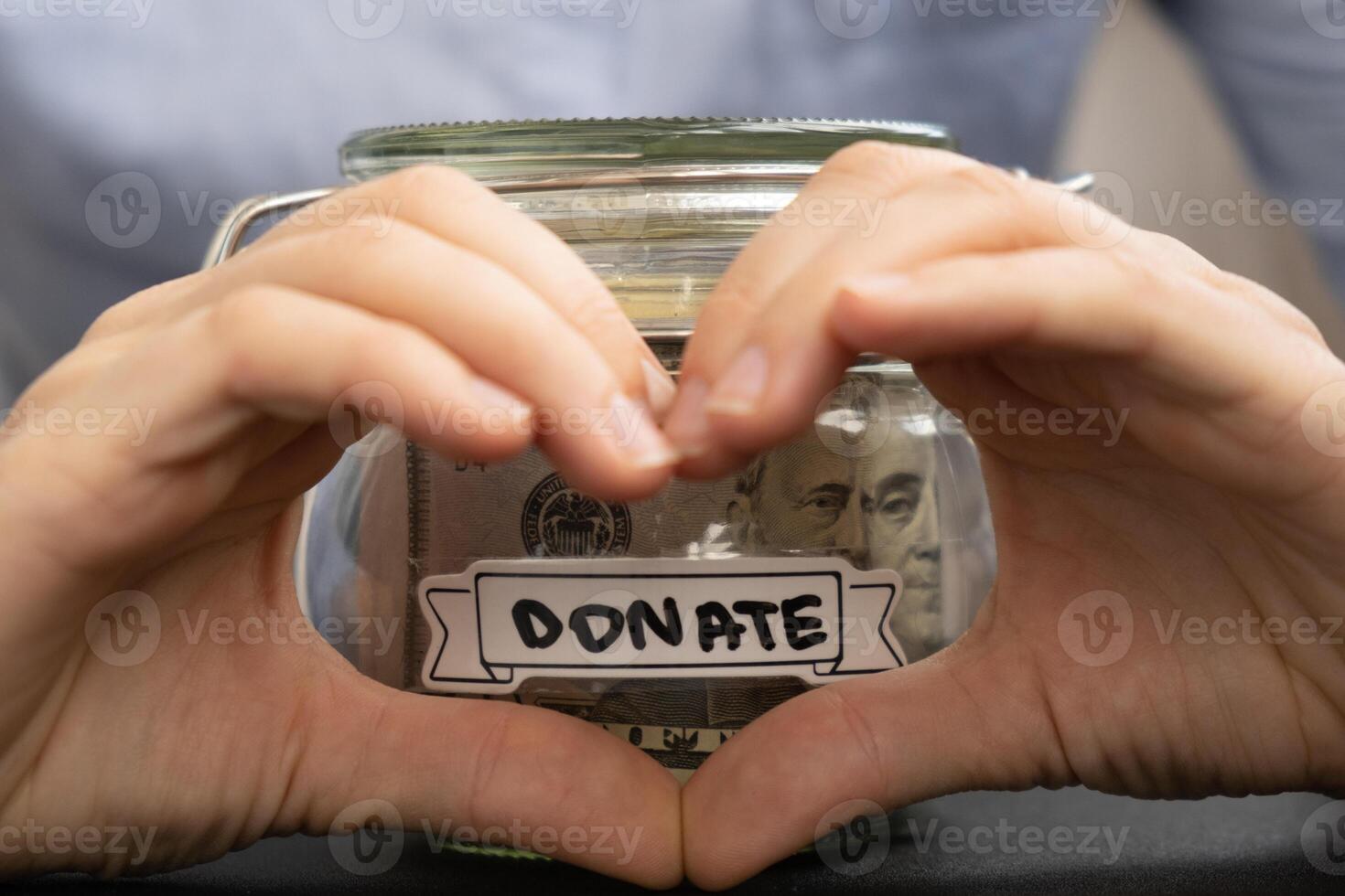Female showing heart sign with hands Saving Money In Glass Jar filled with Dollars banknotes. DONATE transcription in front of jar. Managing personal finances extra income for future insecurity background photo