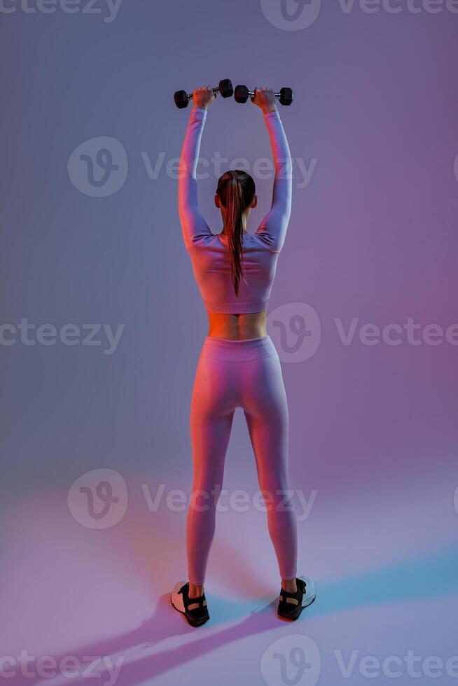 Back view of fitness woman doing exercises with dumbbells on studio background with colored filter photo