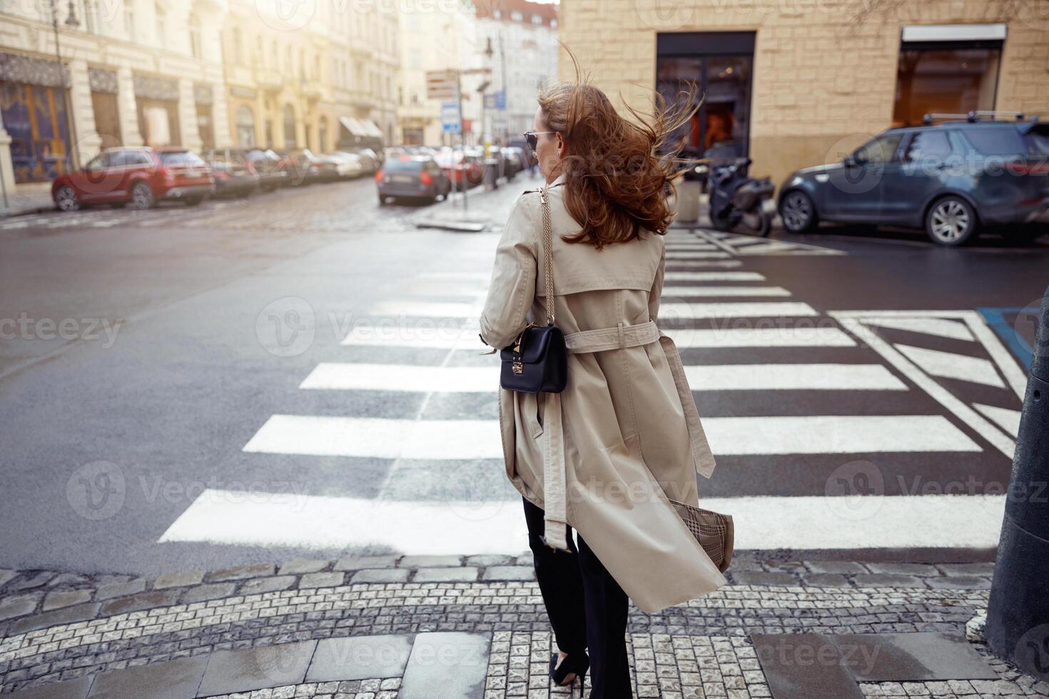 Back view of woman in trendy outfits crosses road against backdrop of city buildings photo