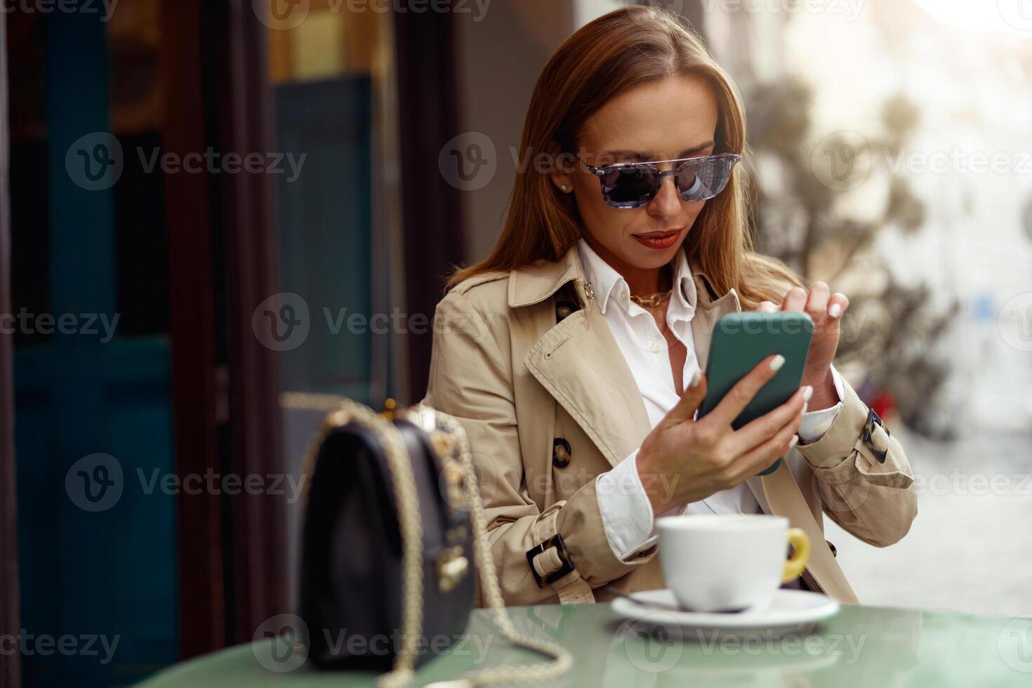 Attractive stylish woman in glasses sitting at cafe terrace and use phone while drinking coffee photo