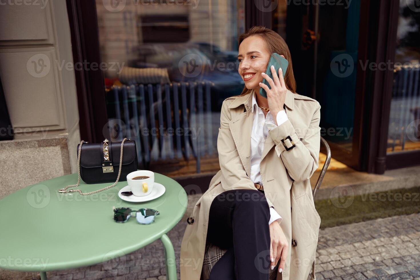 sonriente elegante mujer sentado a café terraza hablando en teléfono mientras Bebiendo café foto