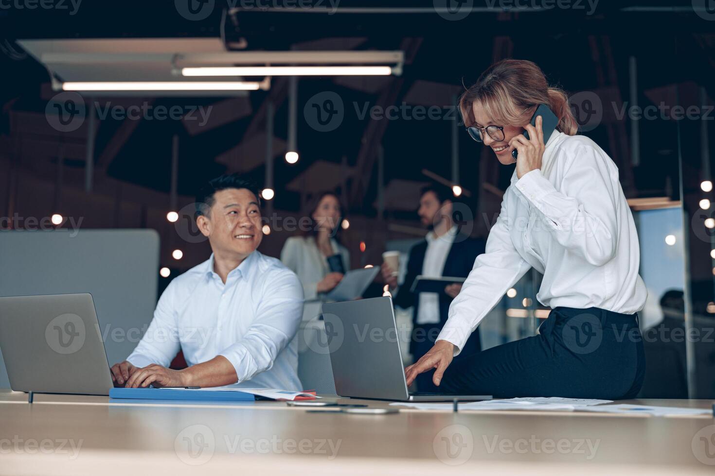 Smiling mature business woman sitting on table and talking phone with client in office photo