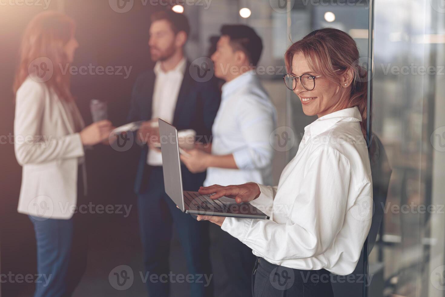 Mature smiling businesswoman at office with group of colleagues on background, working on laptop photo