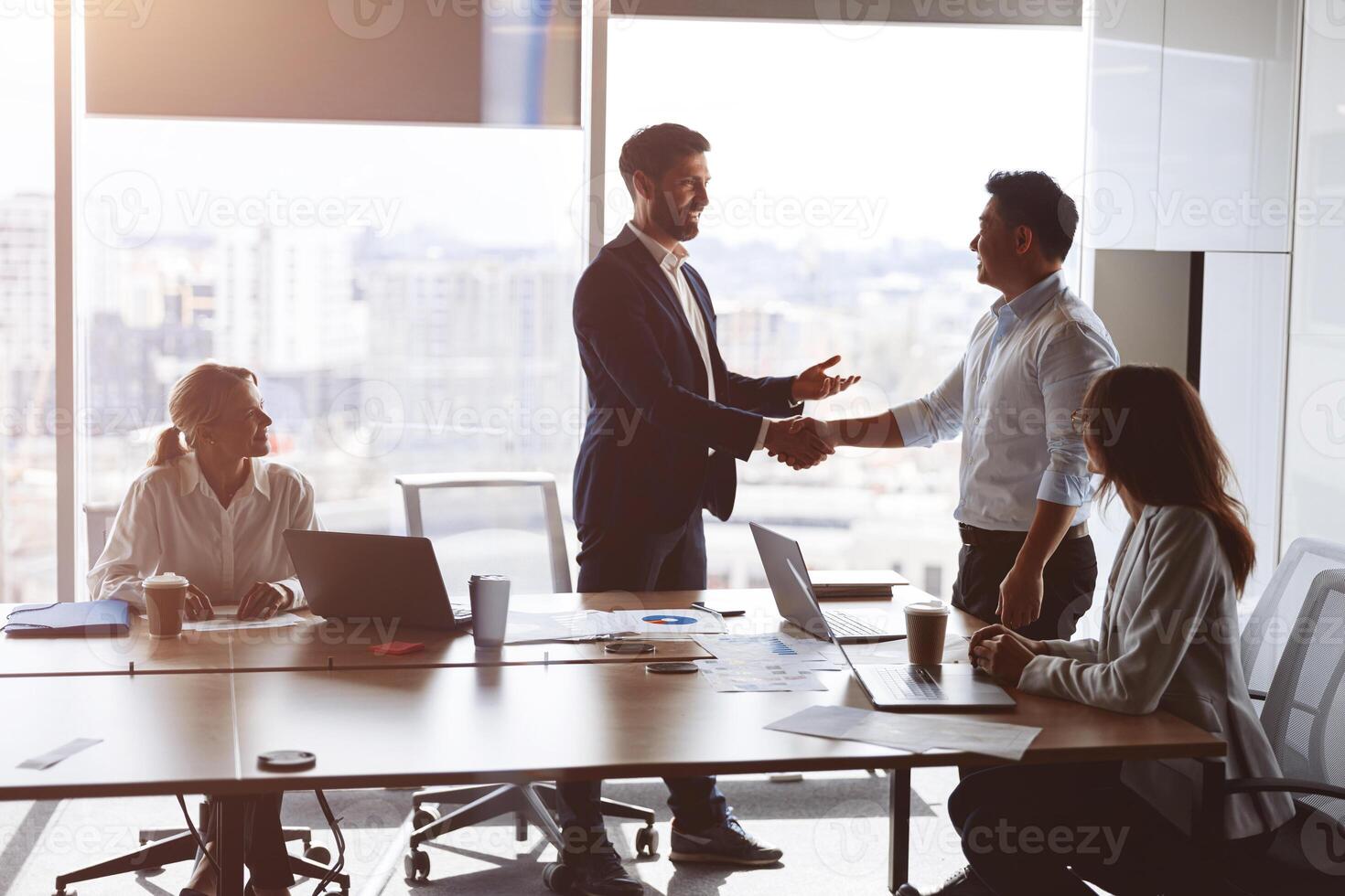 view of confident business partners shaking hands while standing at panoramic window of boardroom photo