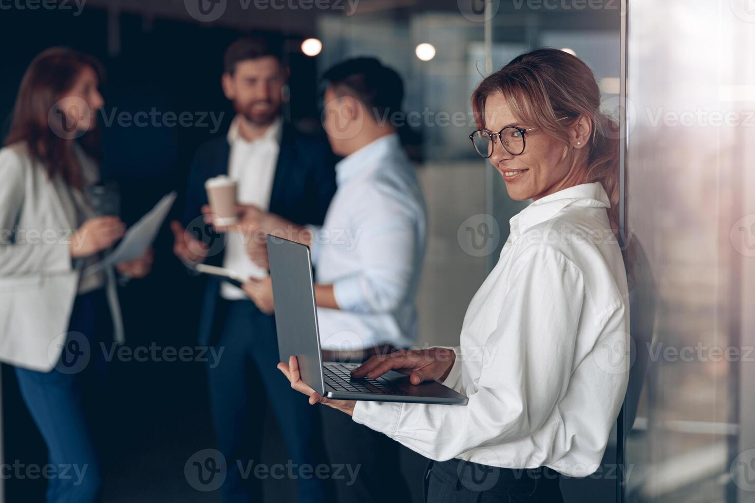 Mature smiling business lady at office with group of colleagues on background, working on laptop photo