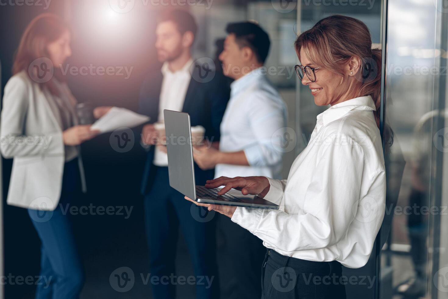 Mature smiling businesswoman at office with group of colleagues on background, working on laptop photo