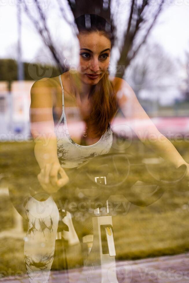 young cute woman in tight sports suit training on exercise bike in gym view through window reflection photo