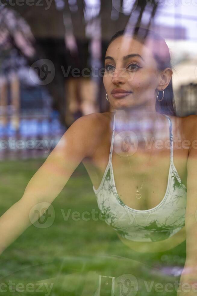 young cute woman in tight sports suit training on exercise bike in gym view through window reflection photo