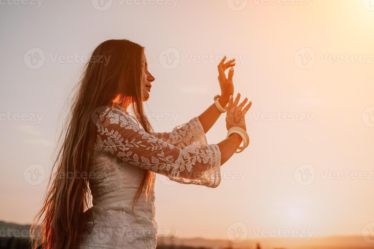 Happy woman in white boho dress on sunset in mountains. Romantic woman with long hair standing with her back on the sunset in nature in summer with open hands. Silhouette. Nature. Sunset. photo