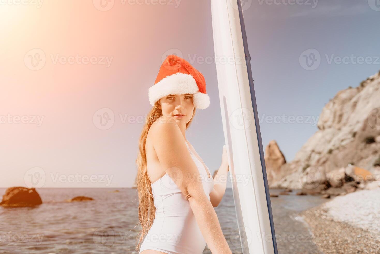 Woman sea sup. Close up portrait of happy young caucasian woman with long hair in Santa hat looking at camera and smiling. Cute woman portrait in a white bikini posing on sup board in the sea photo