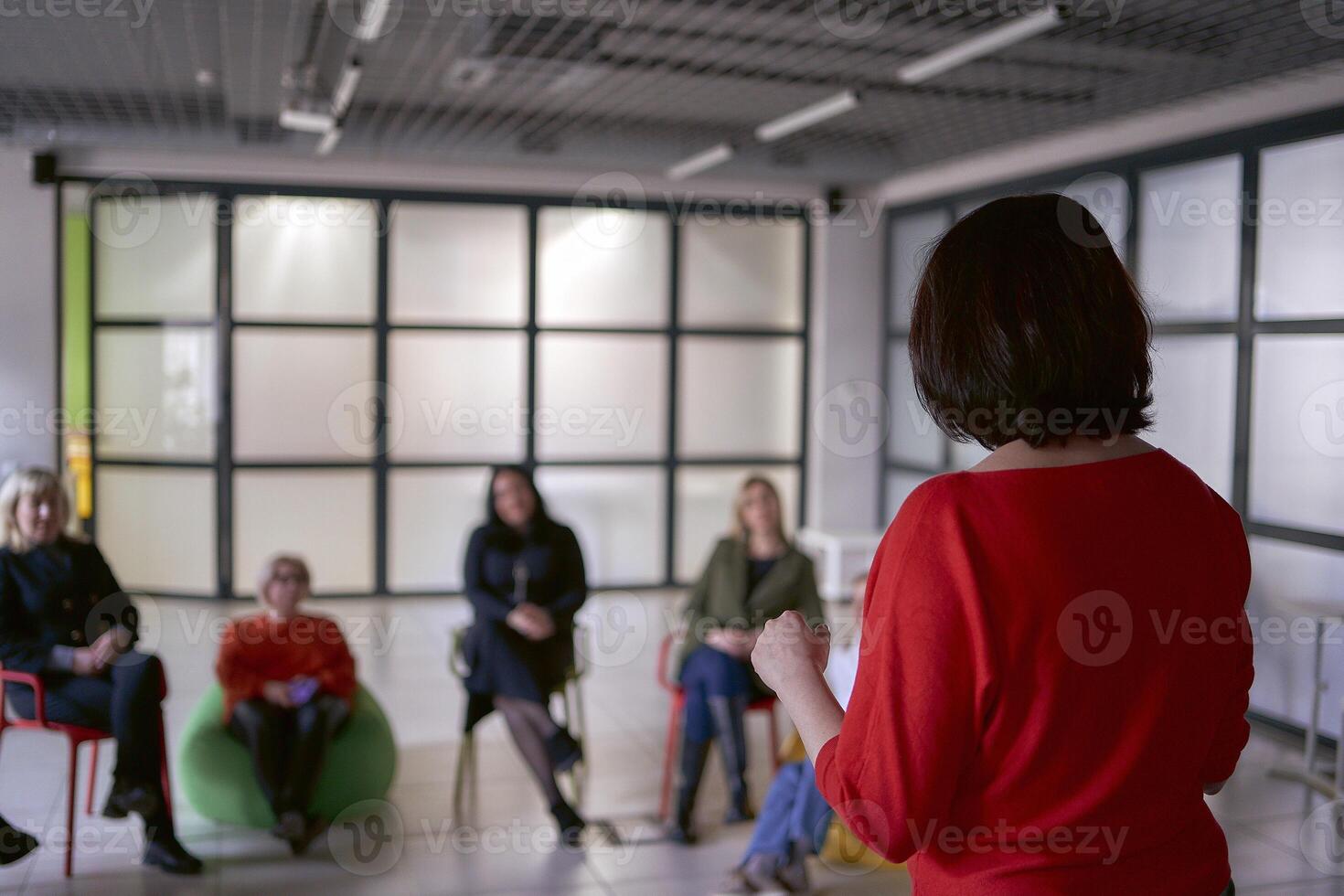 a woman makes a speech on stage, colleagues listen to her speech and sit on chairs and ottomans in the hall photo