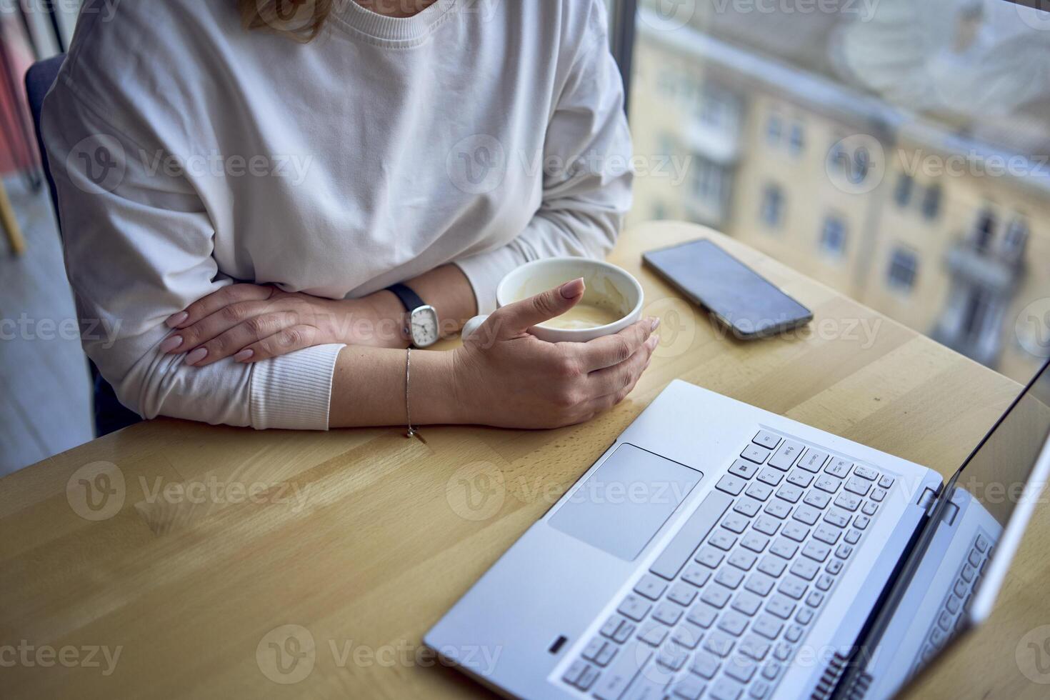 middle age woman with blond hair drinks coffee and works at a laptop in a coworking space with panoramic windows and a view of the city from above, details laptop, coffee, table photo