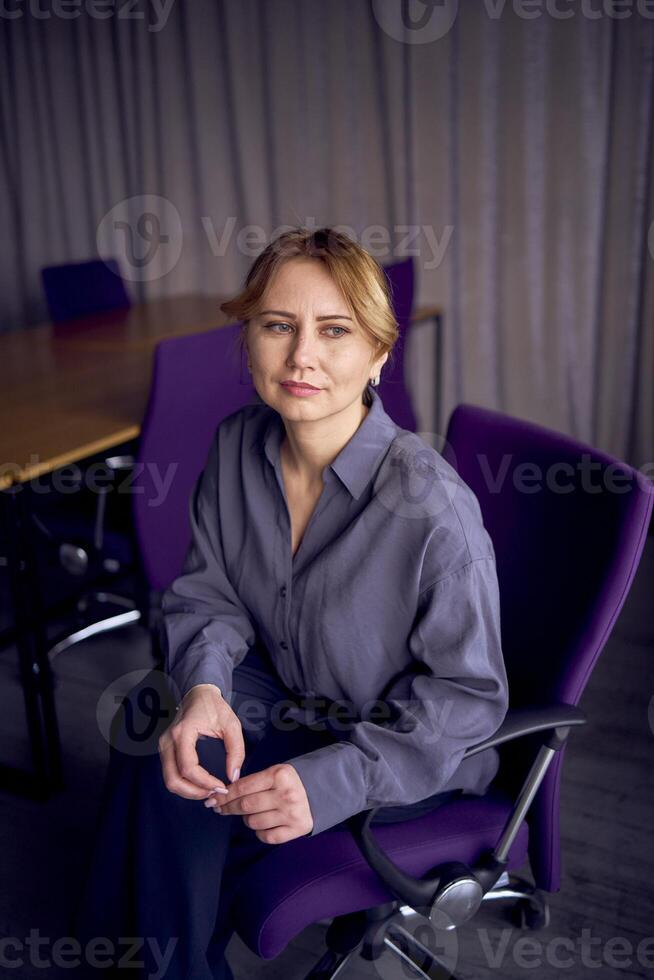 beautiful middle age business woman wearing gray shirt, wide leg pants abd black stilettos sit in purple office chair in modern workspace photo