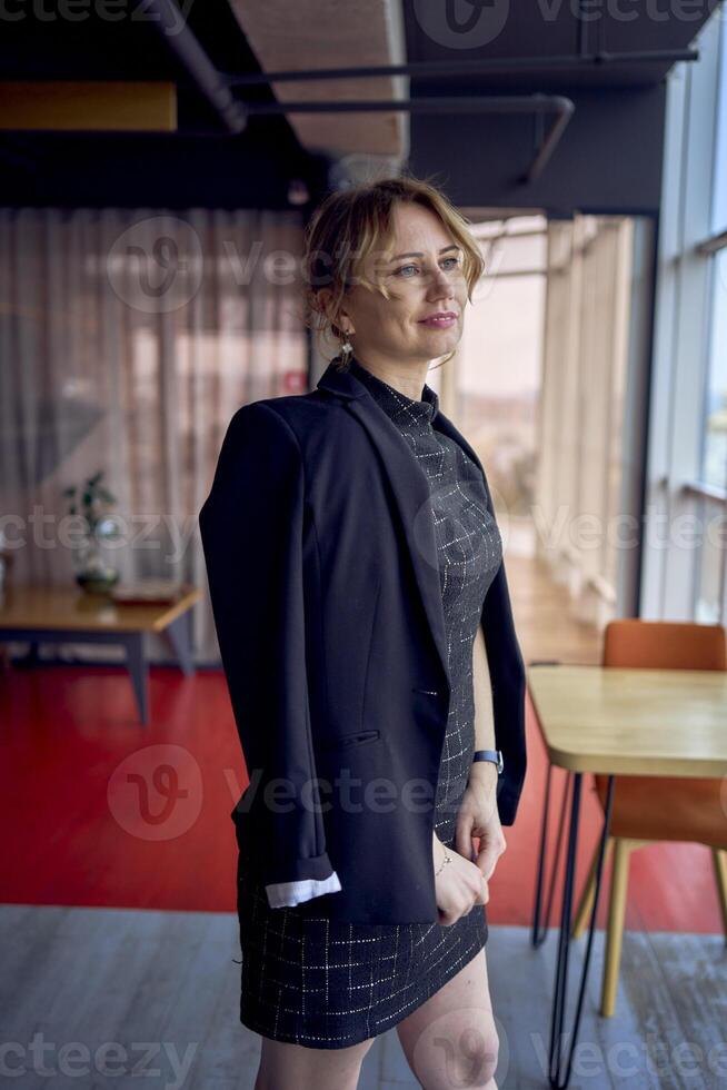 attractive woman in a black business style mini dress throws a jacket over her shoulders in a modern coworking space with panoramic windows and a black ceiling photo