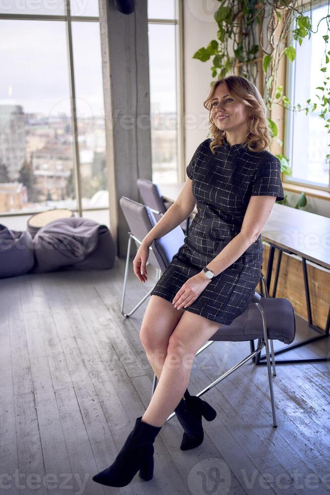Cheerful Young Woman Sitting on a Chair by Large Windows in a Bright Modern Room photo