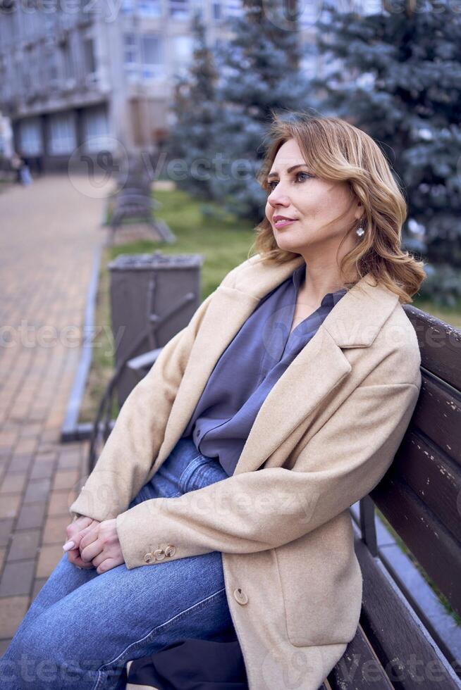 young adult woman in jeans and sand coat sitting on a bench on a spring day, modern architecture behind photo