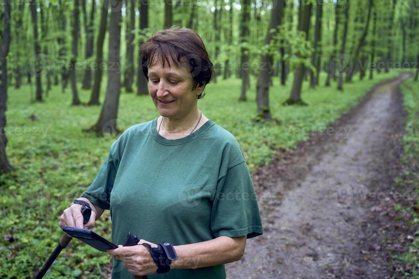 mayor mujer haciendo nórdico caminando con palos en primavera bosque interruptores podcast en teléfono foto