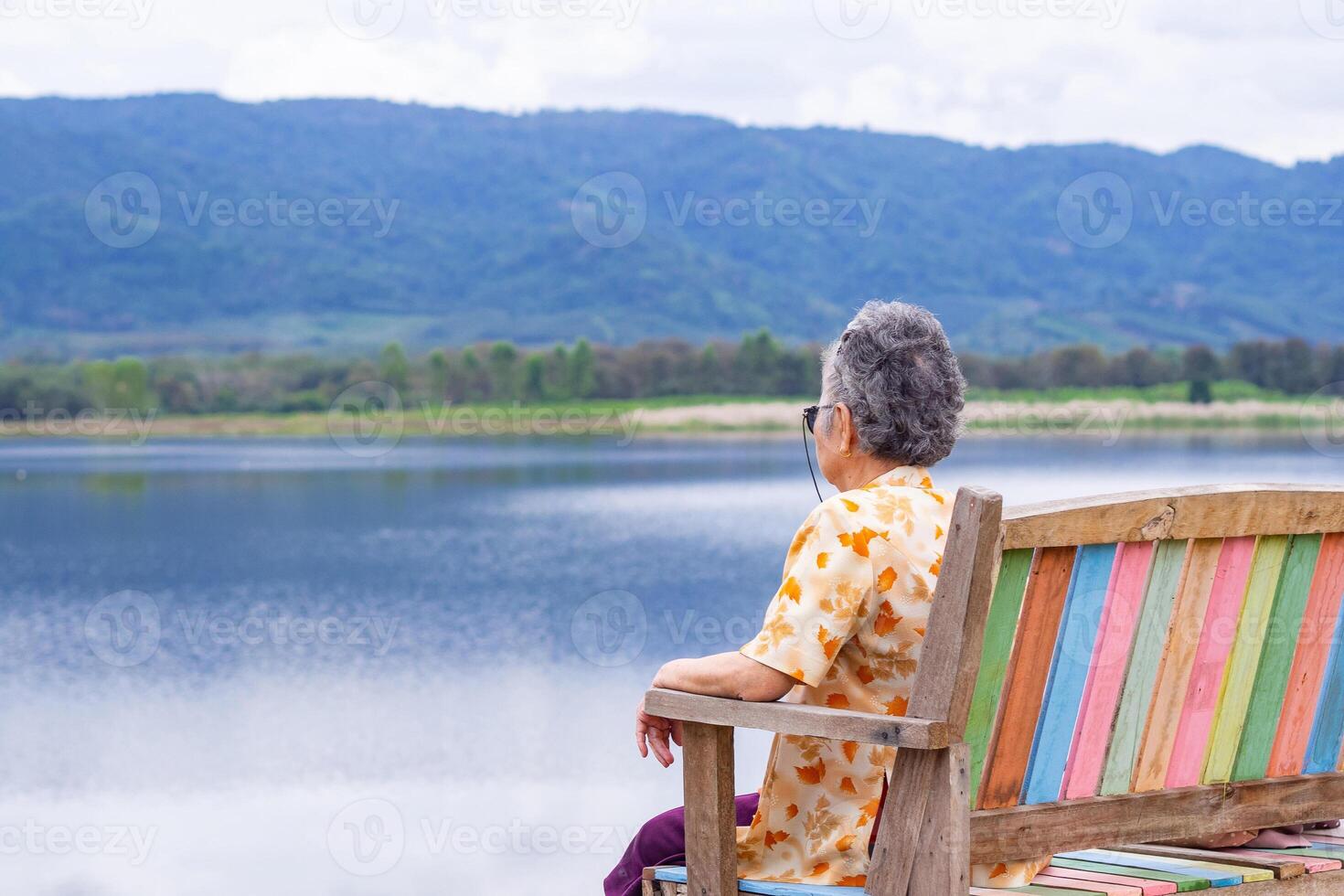 Back view of a senior woman looking to the lake and mountains while sitting on a wooden chair side of the lake. Concept of aged people and relaxation photo