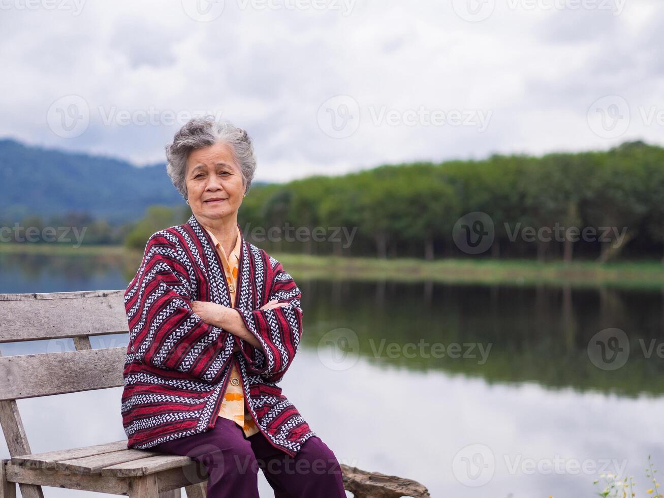 retrato de un mayor mujer sonriente, brazos cruzado y mirando a el cámara mientras sentado lado el lago. espacio para texto. concepto de Envejecido personas y relajación foto