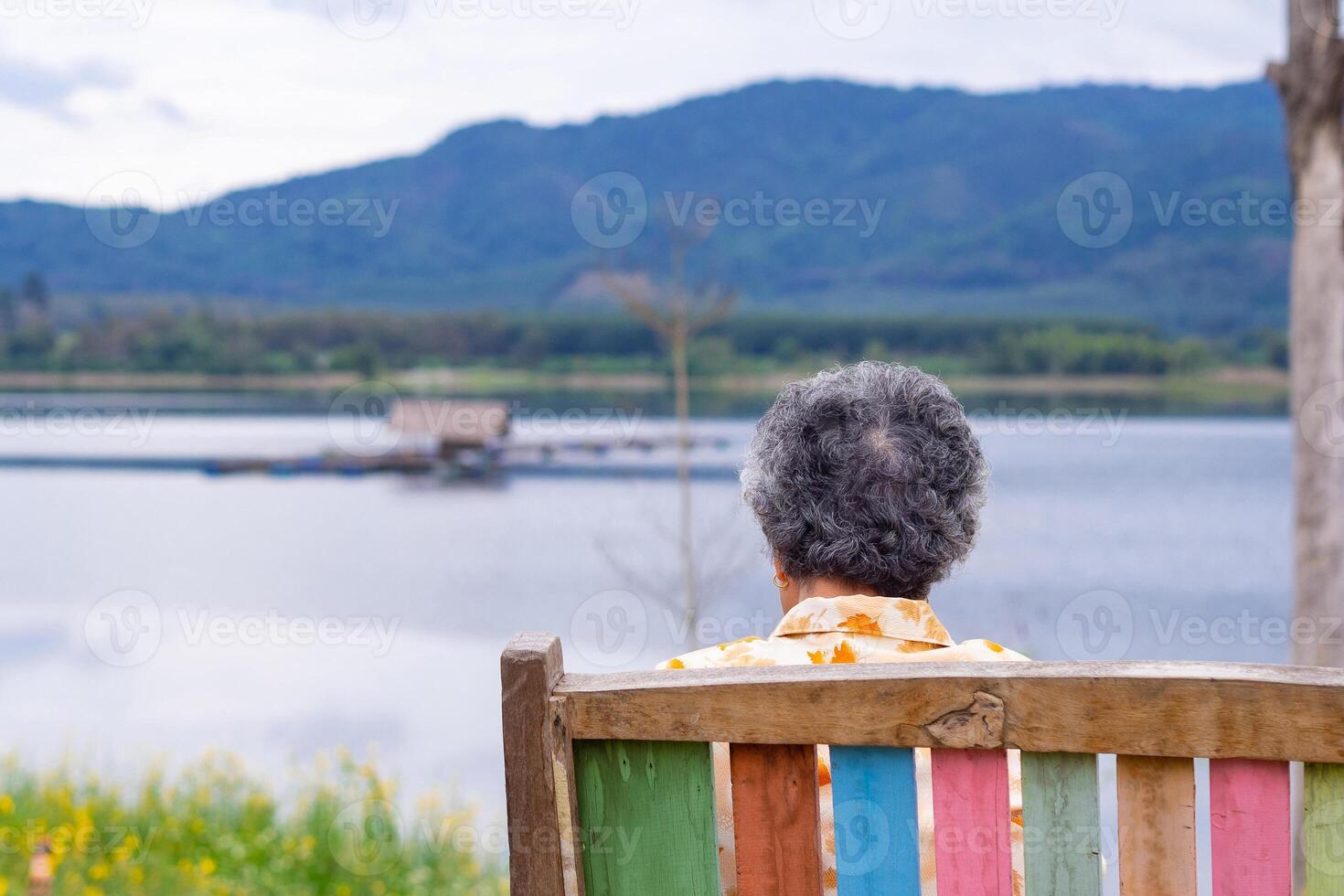Back view of a senior woman looking to the lake and mountains while sitting on a wooden chair side of the lake. Concept of aged people and relaxation photo